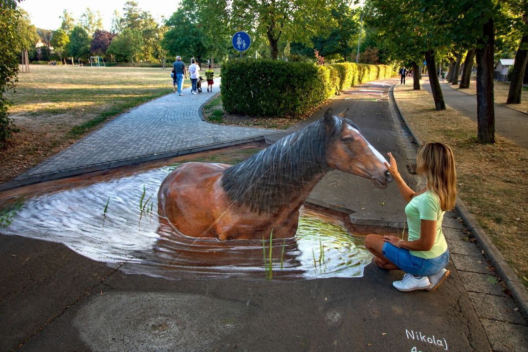 3D street art by Nikolaj Arndt in Neustadt, Germany, depicting a realistic horse standing in a shallow pond, creating an illusion that the horse is emerging from the water onto the pathway. The artist’s detailed work with shadows and reflections gives the horse a lifelike appearance. A woman sits beside the artwork, reaching out to touch the horse’s face, adding to the interactive illusion and highlighting the depth and realism of the piece within the park setting.