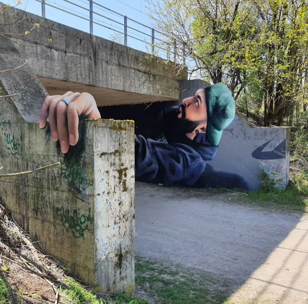 A captivating 3D mural titled Out of the Tunnel by Cosimo Cheone Caiffa in Nerviano, Milan, Italy. The artwork depicts a bearded man in a green cap and hoodie appearing to crawl out of a concrete underpass, with his hand gripping the edge of the wall. The hyper-realistic details and clever use of the structure's dimensions create a stunning optical illusion, making it seem as if the man is breaking through the urban environment. The mural's interaction with its surroundings adds a dynamic and immersive quality to the piece.