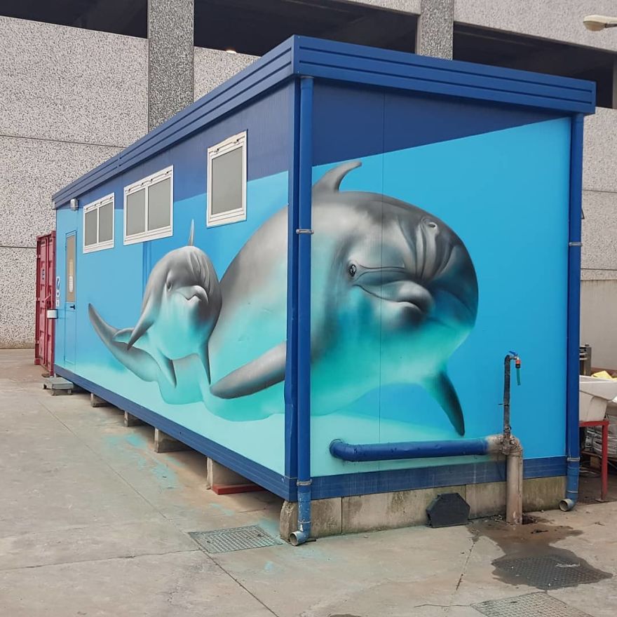 A vibrant and lifelike mural by Cosimo Cheone Caiffa featuring two dolphins painted on a portable container. The artwork uses realistic shading and aquatic tones to create the illusion of dolphins swimming gracefully in a bright blue underwater scene. The surrounding elements of the container, such as pipes and edges, are seamlessly incorporated into the design, enhancing the sense of depth and motion. This imaginative transformation of an industrial structure into a marine-themed artwork highlights Cheone’s talent for bringing life to unexpected urban surfaces.