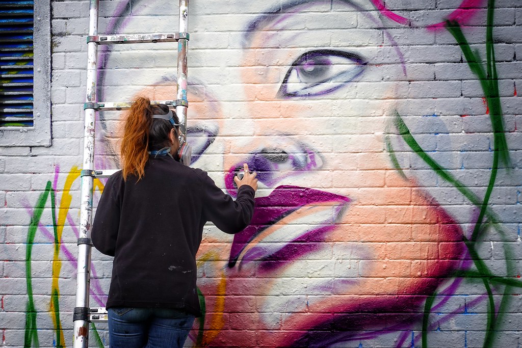 A young woman in a black top and blue jeans and with dark hair and a ginger pony tail stands by a ladder up against a wall. She is holding a spray can in her right hand, adding detail to a large painting of a woman's face which is mostly outline with only the lower part of the face filled in