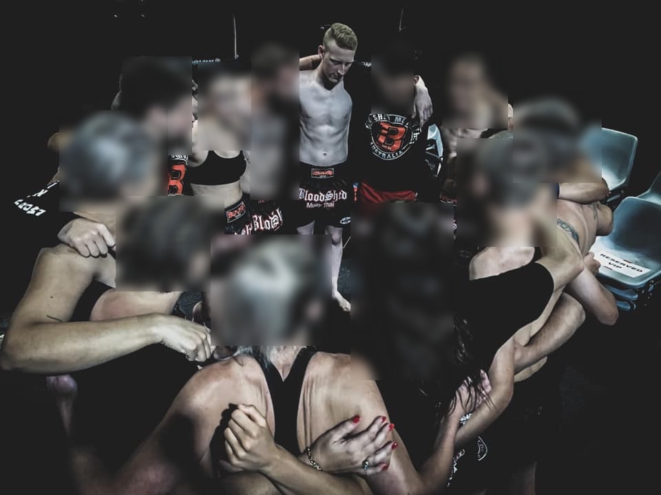 A group photo of scrawny Gabe in a huddle with twelve other people in Muay Thai clothes. His pale skin looks grey in the low light.