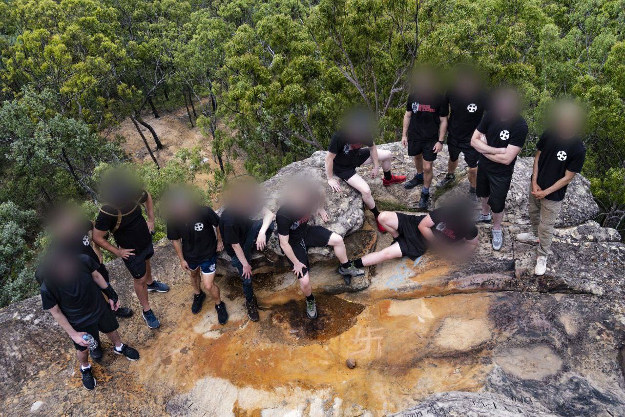 A larger version of the same photo showing twelve neo-Nazis dressed in black posing for a photograph at the top of White Rock in Ipswich. They have carved a swastika into the rock, desecrating this sacred site.