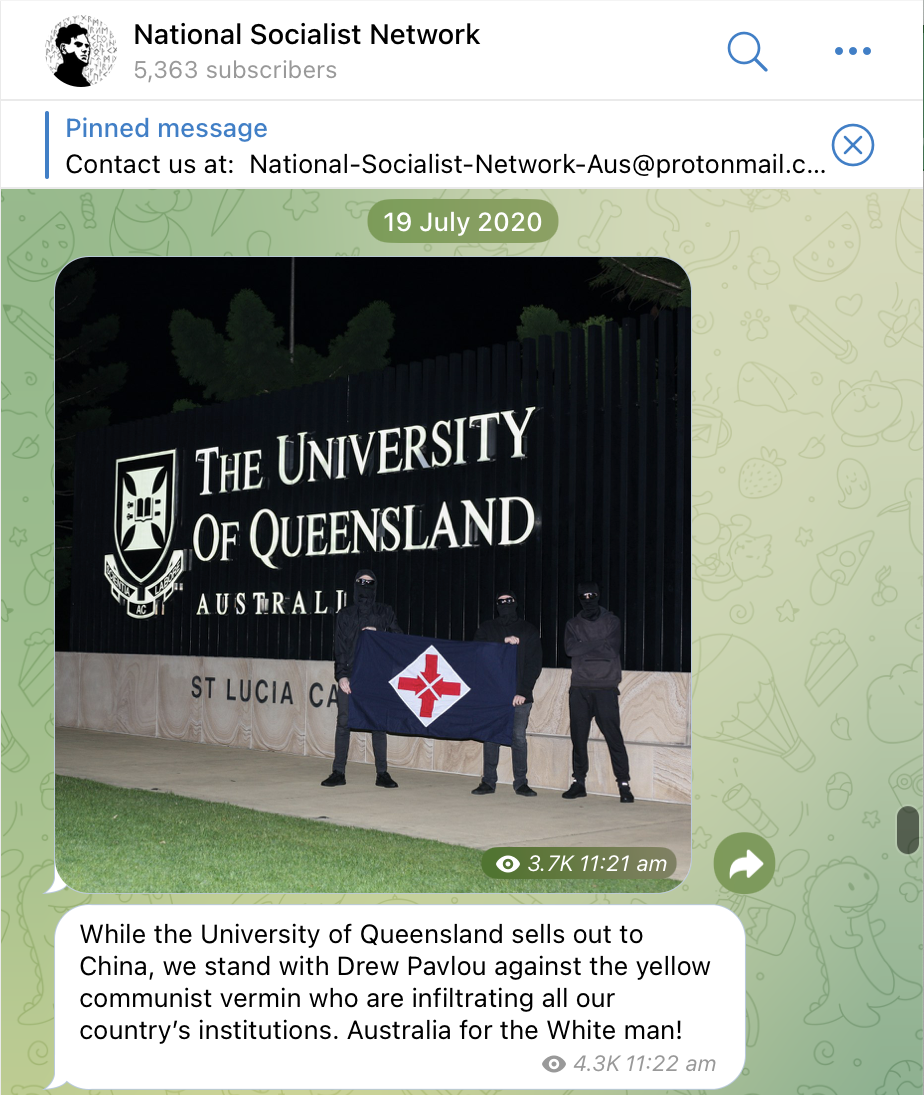 A screenshot of the National Socialist Network Telegram channel of a post dated 19 July 2020. Three neo Nazis stand in front of The University of Queensland St Lucia campus sign. They are wearing all black and black balaclavas. Two of them are holding a National Socialist Network flag, which is an altered swastika. Their caption reads, While the University of Queensland sells out to China, we stand with Drew Pavlou against the yellow communist vermin who are infiltrating all our countrys institutions. Australia for the White man.