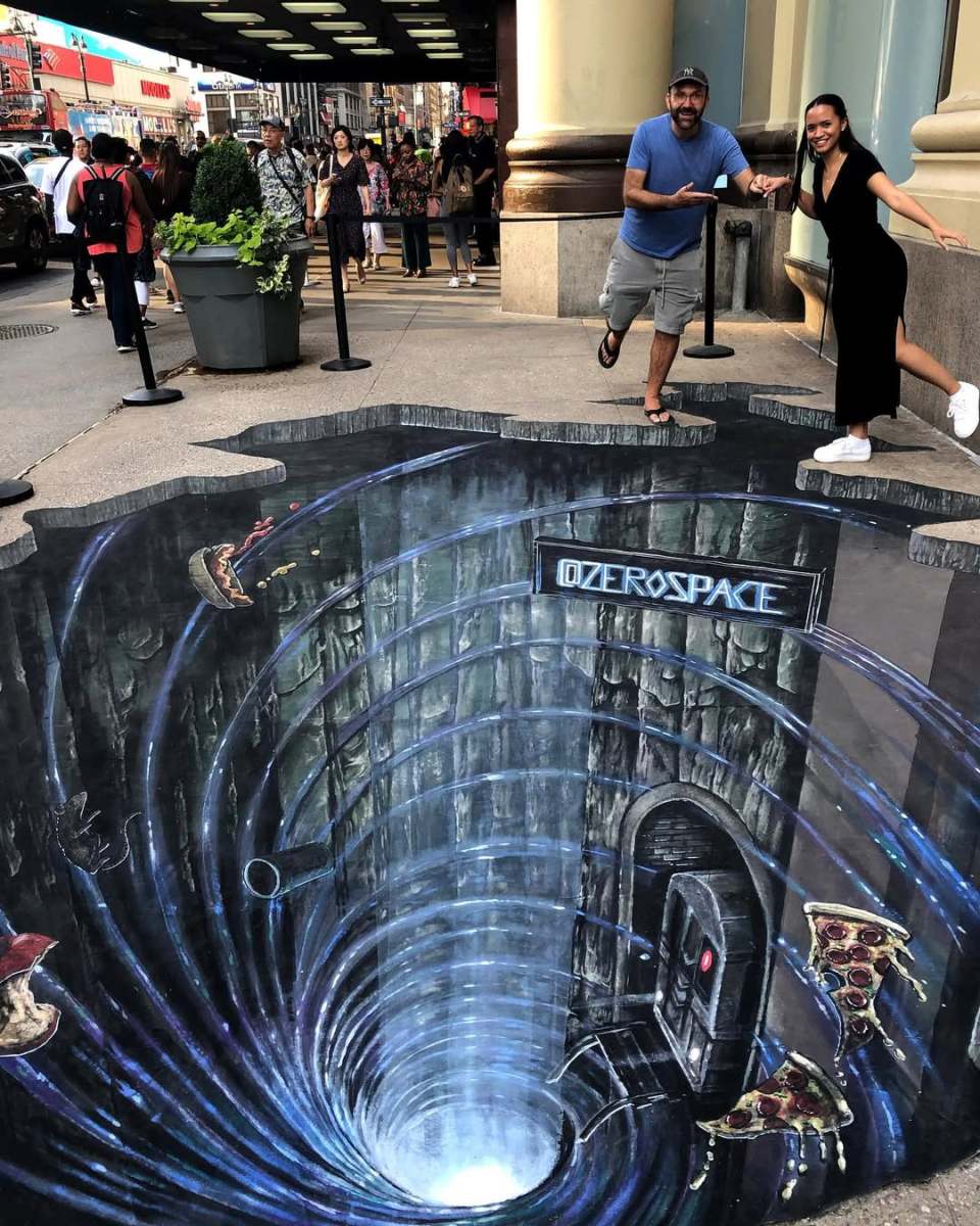 A thrilling 3D street art illusion by Joe and Max, featuring a deep vortex labeled "ZeroSpace," with swirling lights and a futuristic doorway at the edge. Slices of pizza and other objects appear to be falling into the abyss, adding a playful touch. Two people pose at the edge, pretending to balance precariously above the chasm, while a bustling city street and curious onlookers serve as the backdrop. This artwork captivates viewers with its blend of humor and immersive design.