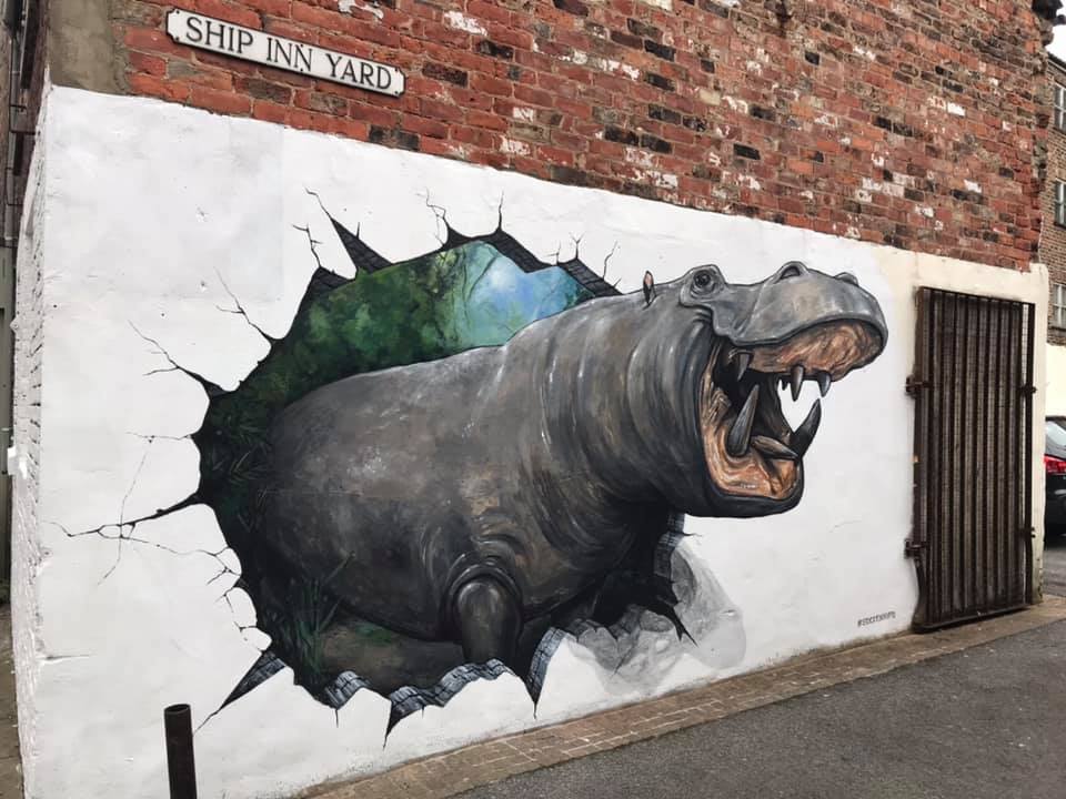 A vivid 3D street art mural by Joe and Max depicting a life-sized hippo bursting through a wall. The artwork gives the impression of the animal emerging from a jungle scene into an urban alleyway, complete with realistic cracks around the wall's edges. Located under a "Ship Inn Yard" street sign, the piece blends humor and hyper-realism, surprising passersby with its striking detail.