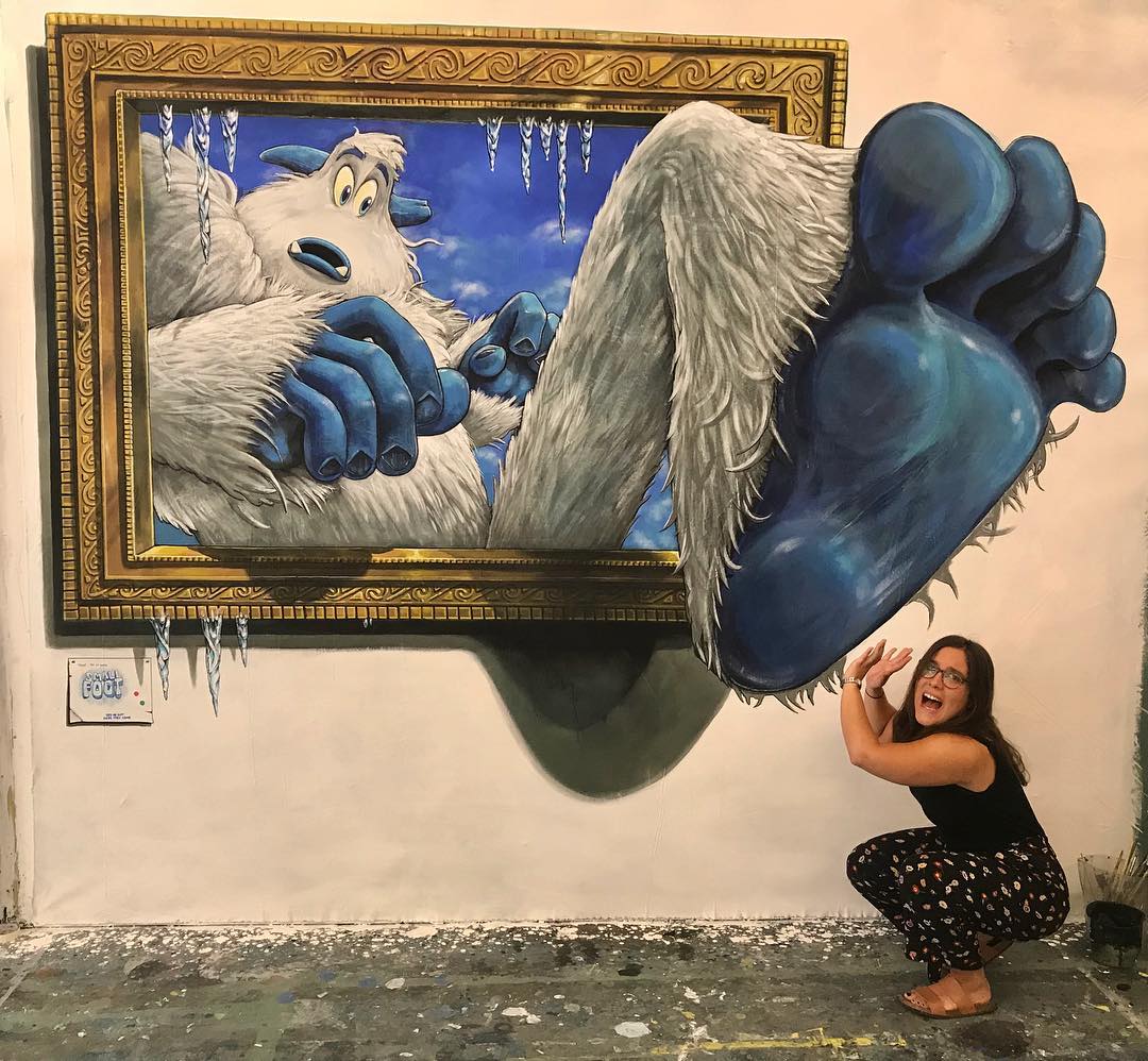 A playful 3D street art mural by Joe and Max featuring a yeti breaking out of a picture frame. The yeti's oversized blue feet appear to extend into the viewer's space, creating a striking illusion of depth. A woman crouches nearby, pretending to shield herself from the enormous feet, adding to the interactive and humorous nature of the artwork. Icicles hanging from the frame enhance the chilly, whimsical atmosphere.