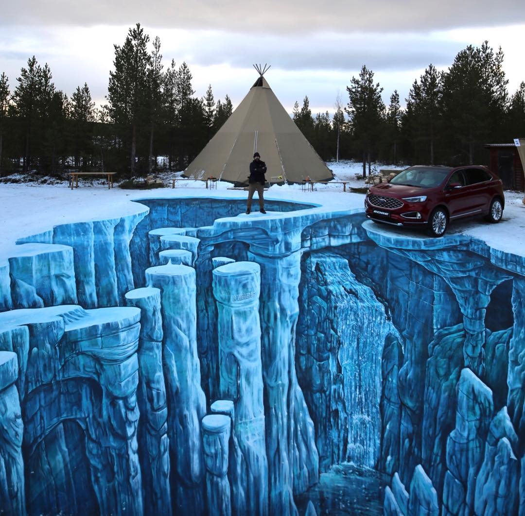 A breathtaking 3D street art installation by Joe and Max, depicting an icy canyon with towering blue pillars and a cascading waterfall. The illusion is painted on snow, with a man standing confidently in the center and a red SUV parked at the edge, as though balanced above the abyss. A teepee in the snowy background adds a serene, wintery atmosphere to this visually stunning and immersive artwork.