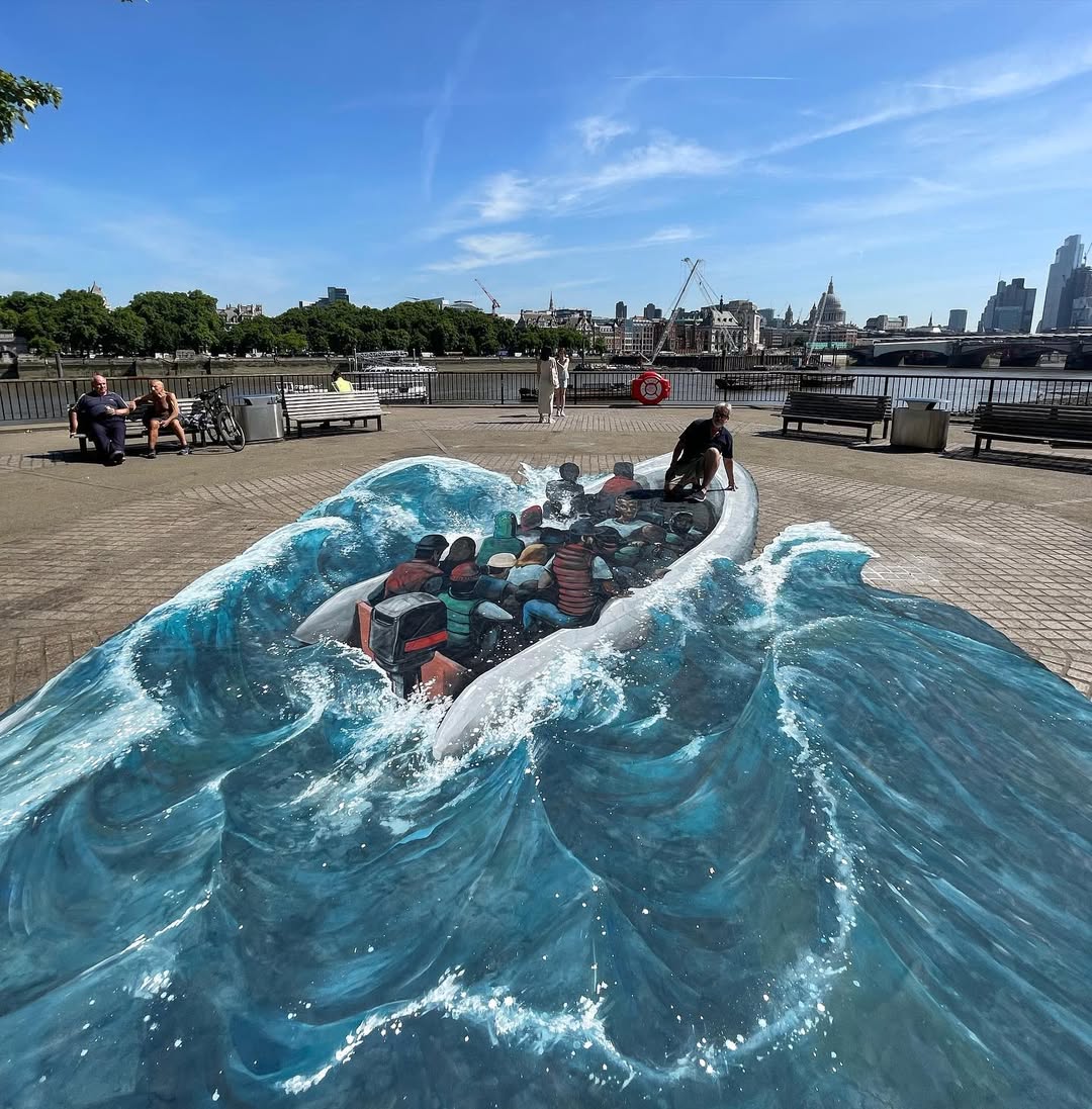 A powerful 3D street art piece by Joe and Max showing a crowded inflatable boat navigating through rough waves, painted directly on a riverside promenade. The realistic artwork captures the sense of motion and urgency as water splashes around the boat. A man interacts with the piece, appearing to climb onto the boat. The London skyline, with iconic landmarks and a sunny sky, forms the backdrop, grounding this thought-provoking artwork in a contemporary urban setting.