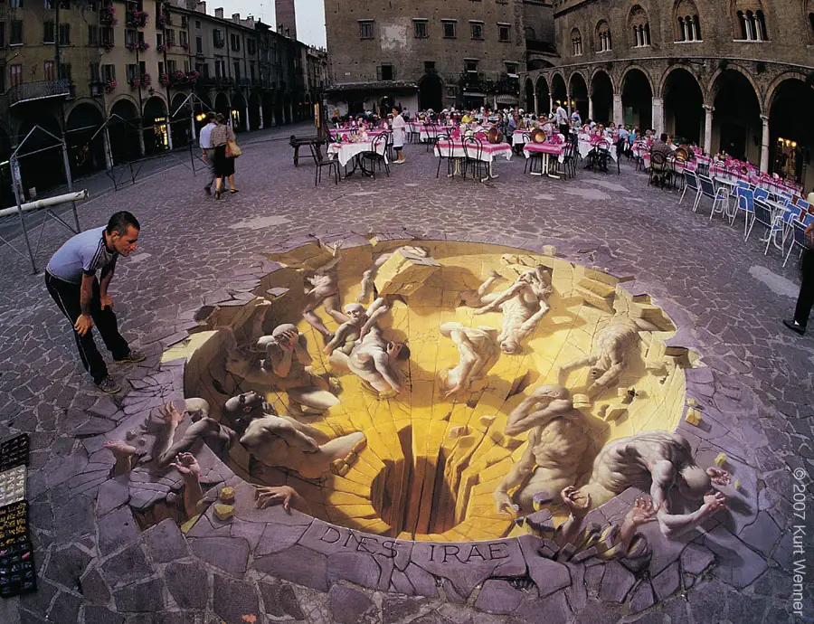 3D pavement art by Kurt Wenner, illustrating a dramatic scene of tormented figures descending into a fiery chasm. The artwork, set in a public square with dining tables in the background, creates a stunning optical illusion of depth, as if the cobblestone ground has cracked open to reveal a pit filled with distressed, human-like figures reaching and writhing in agony. A man leans in to observe the illusion closely, enhancing the interactive effect. The phrase 'Dies Irae' (Day of Wrath) is inscribed on the ground, adding a thematic layer to the powerful, immersive artwork.