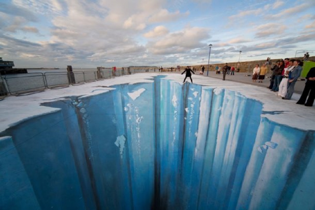 3D pavement art by Julian Beever, creating an illusion of a deep icy chasm on a flat surface near a waterfront. The artwork depicts a massive, vertical drop with sharp, frozen cliffs, as if the ground has cracked open to reveal an icy abyss. A person stands precariously at the edge of the artwork, adding a sense of danger and realism to the scene. Spectators watch from a safe distance, enhancing the interactive and immersive nature of Beever's illusion.