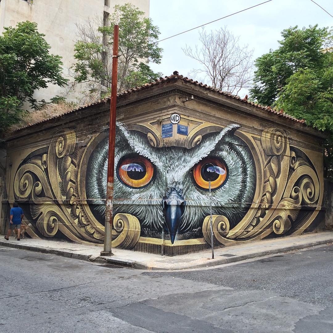 Mural titled ‘Knowledge speaks – Wisdom listens’ by artist WD (Wilddrawing) in Athens, Greece. The artwork covers the corner of a building, transforming it into a massive, realistic owl face with intense, bright orange eyes that seem to follow passersby. Intricate gold and brown decorative elements surround the owl, blending classical architectural motifs with street art. The mural’s detailed textures and striking gaze create a powerful visual effect, symbolizing wisdom and knowledge in the heart of the city. A pedestrian walking by gives a sense of scale to the imposing owl mural.