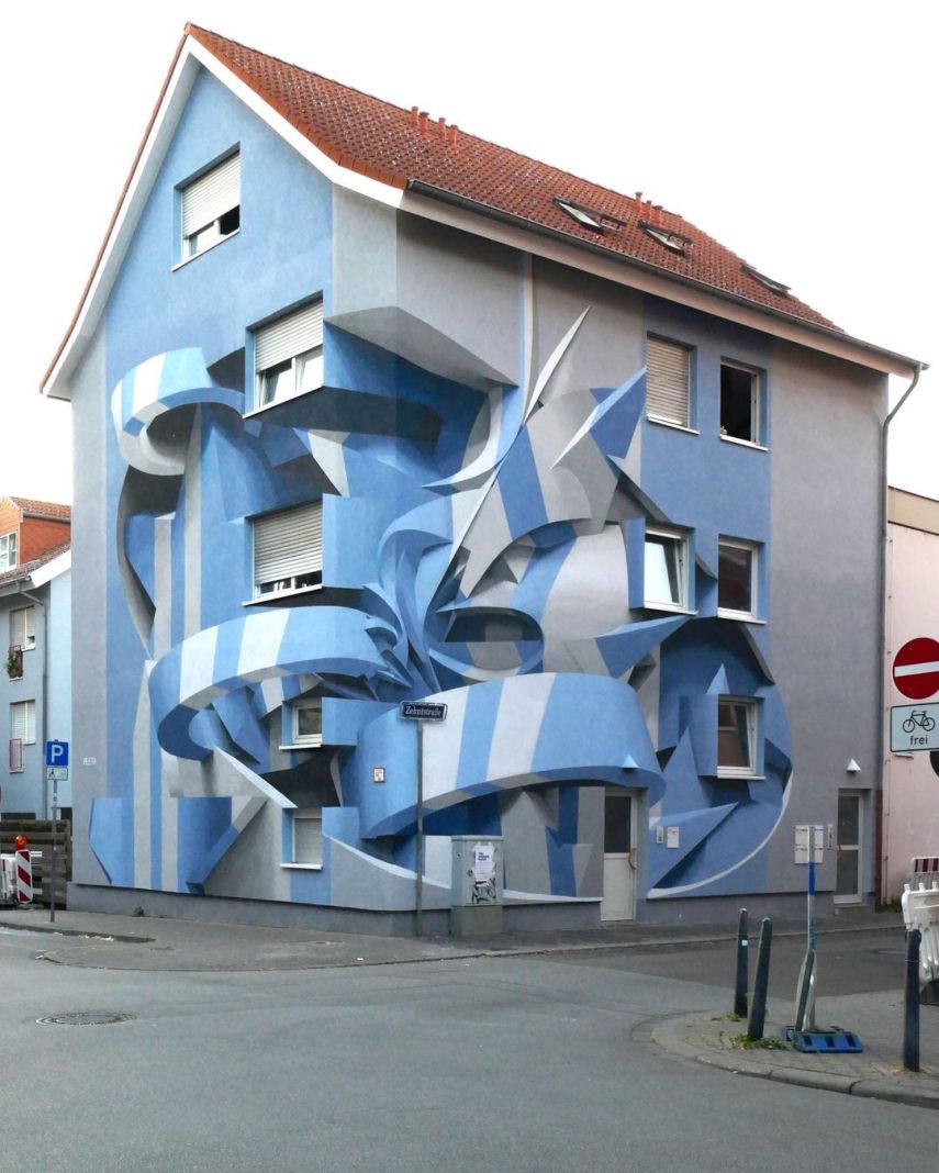 3D mural by artist Peeta on a residential building in Mannheim, Germany. The artwork uses shades of blue and white to create the illusion of twisting, ribbon-like shapes that appear to wrap around and cut into the building’s facade. The intricate design gives the impression of depth and movement, transforming the flat walls into a dynamic, sculptural form. The realistic shadows and contours enhance the optical illusion, making the structure appear as if it’s deconstructed and reformed into abstract shapes. The surrounding street and signage add context to the urban setting.