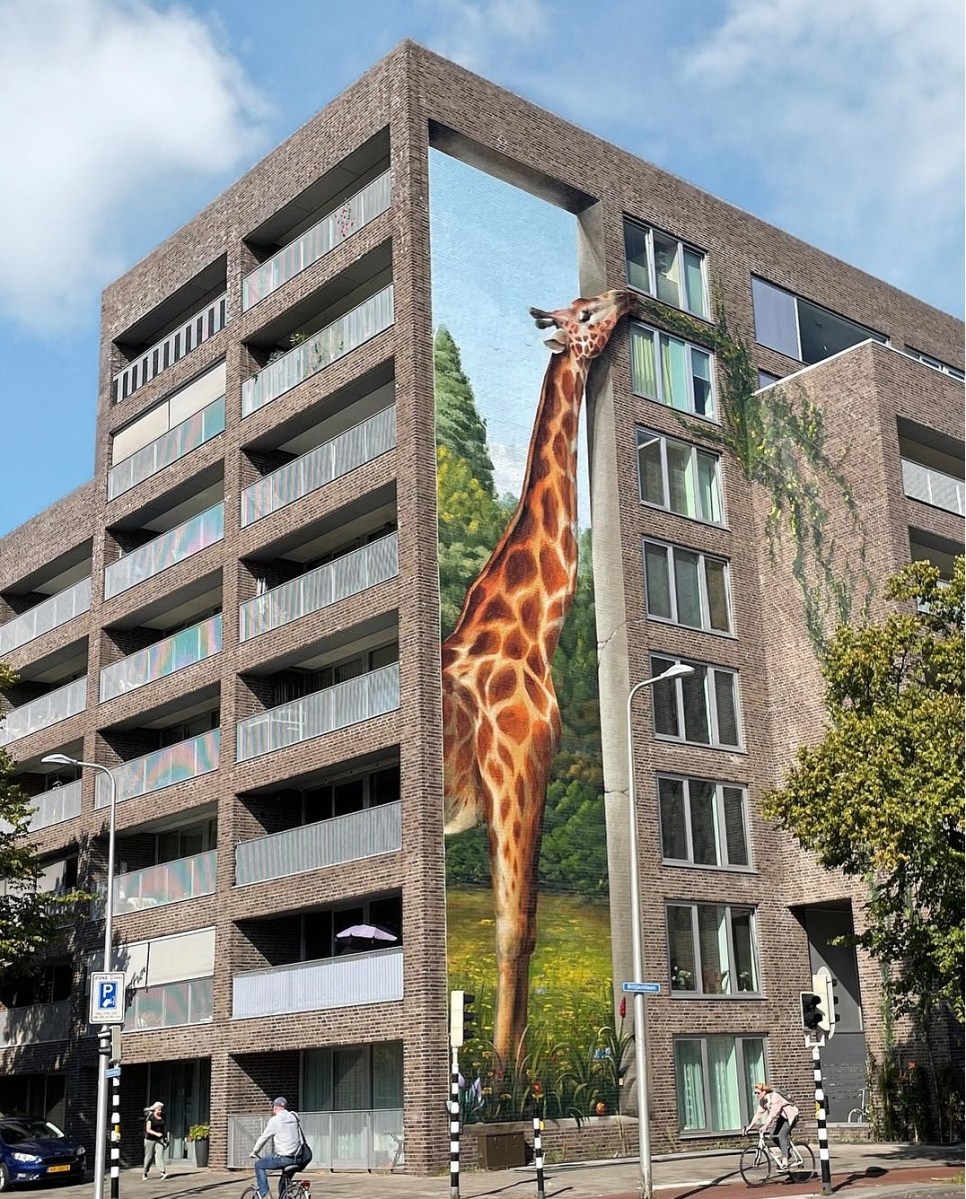 Mural titled 'Giraffe Eating the Plants' in Utrecht, Netherlands, depicting a realistic giraffe reaching up to nibble on plants growing on the balcony of a residential building. The artwork covers the corner of the building, creating an optical illusion that integrates the giraffe seamlessly with the architectural structure. The background of the mural shows lush greenery, blending urban and natural elements in a playful way. Cyclists and pedestrians in the foreground add to the urban setting, while the towering giraffe mural adds whimsy and charm to the neighborhood.