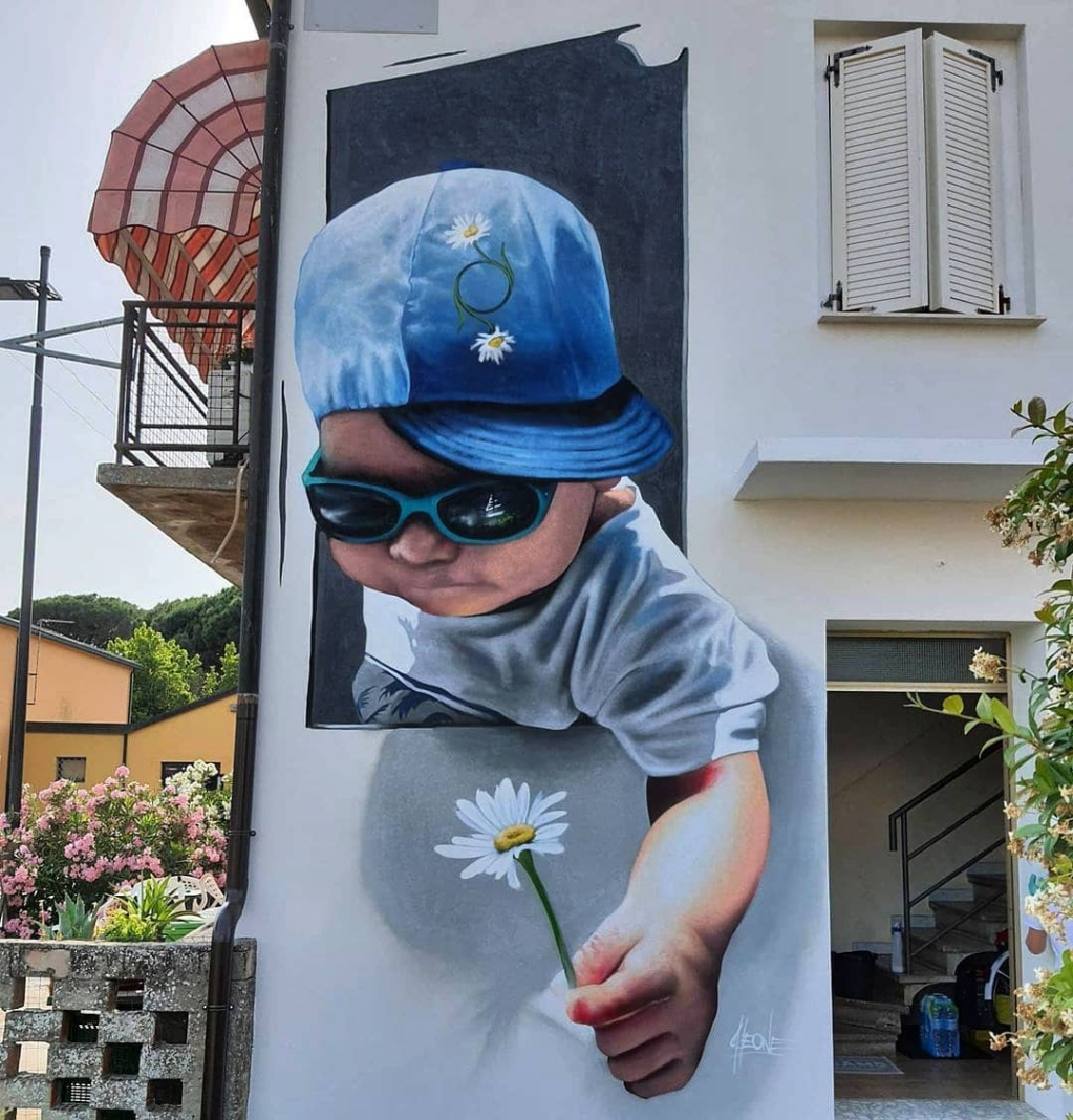 A hyper-realistic mural by 3D street artist Cheone in Porto Viro, Italy, depicting a young child wearing a blue cap adorned with small daisies and reflective sunglasses. The child reaches out of the painted frame, holding a delicate white daisy, creating an interactive 3D illusion. The vibrant colors and lifelike details make the artwork blend seamlessly with the surrounding architecture and natural greenery.