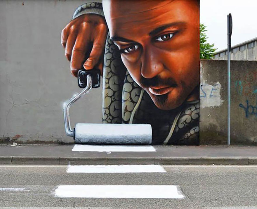 A brilliant 3D mural by Cosimo Cheone Caiffa in Trezzano sul Naviglio, Milan, Italy, depicting a man holding a paint roller. The artwork cleverly interacts with its surroundings as the painted roller appears to extend onto the road, creating a striking illusion of the man painting the pedestrian crossing. The hyper-realistic style, combined with the integration of the urban environment, blurs the line between art and reality, showcasing Cheone's mastery in creating immersive, site-specific street art.