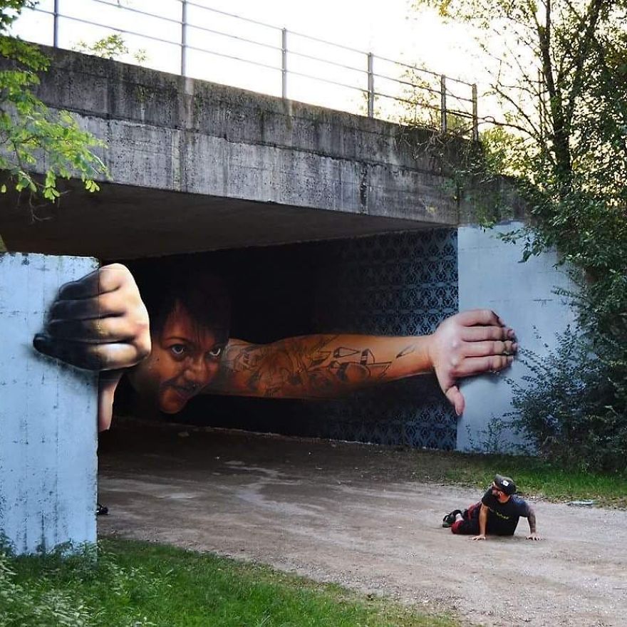 A striking 3D mural collaboration by Cosimo Cheone Caiffa and Mor Pavone in Nerviano, Italy, featuring a powerful figure emerging from under a concrete bridge. The figure, depicted with tattooed arms outstretched, appears to be gripping the sides of the bridge, creating a dramatic illusion of breaking free. A child sits on the ground in front of the mural, further enhancing the interaction between the artwork and its surroundings. The meticulous details in the tattoos, facial expression, and hand positioning add depth and realism to this captivating piece of street art.