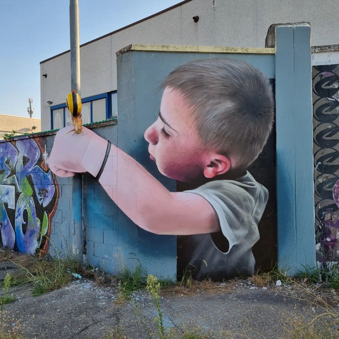 A hyper-realistic 3D mural by Cosimo Cheone Caiffa featuring a young boy extending his arm to touch a yellow-black bollard, seamlessly integrating the real-world object into the artwork. The boy's delicate features, rosy cheeks, and focused gaze create a tender and lifelike scene that blurs the boundary between the mural and its surroundings. The artwork's interplay between painted elements and physical objects showcases Cheone's mastery in creating immersive and interactive street art.