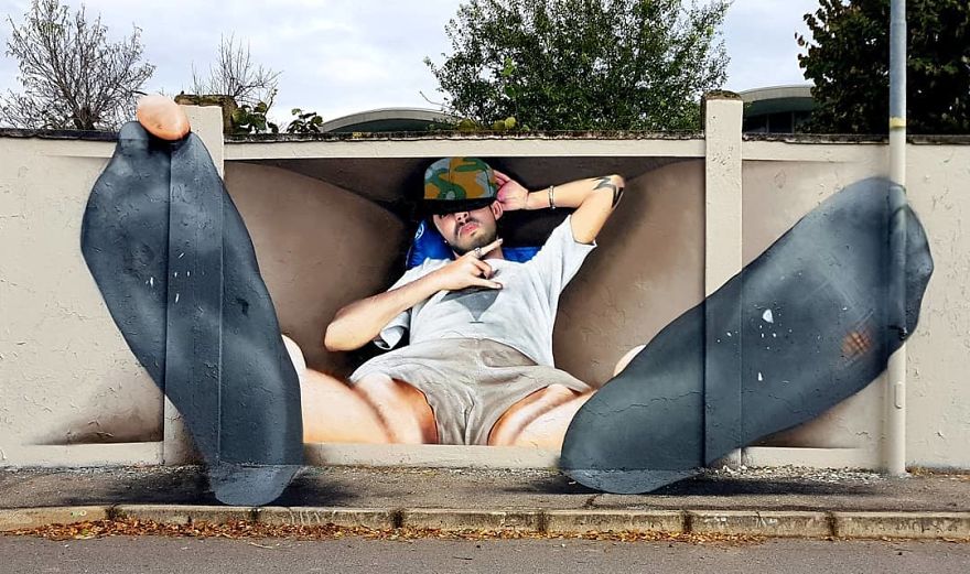 A humorous and creative 3D mural by Cosimo Cheone Caiffa depicting a relaxed man lounging in a confined space painted on a wall. The man, wearing a camouflage cap and casual attire, leans back with his feet extended outwards, creating a striking illusion that his legs are protruding into the viewer's space. The oversized feet, complete with realistic textures and shadows, amplify the playful nature of the artwork. This clever piece blurs the line between two-dimensional art and reality, showcasing Cheone's exceptional skill in perspective and trompe-l'œil techniques.