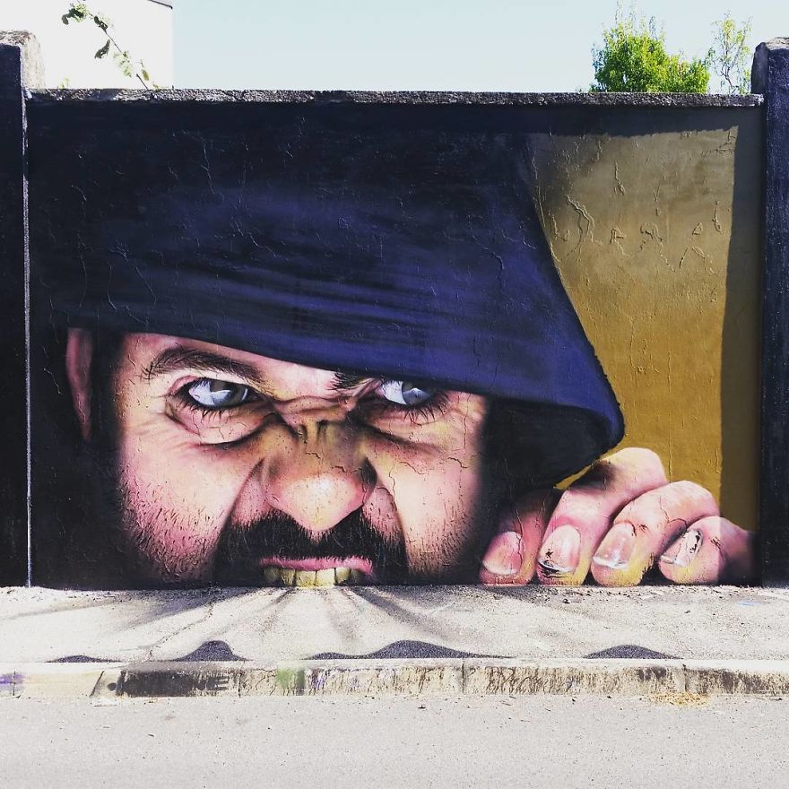 A powerful 3D mural by Cosimo Cheone Caiffa depicting a man with an intense expression, wearing a dark hood. The man appears to be gripping the edge of the wall with his fingers, his teeth clenched and his eyes glaring with a mix of determination and ferocity. The hyper-realistic details in his face, including the texture of his skin and the dirt under his nails, create a striking and lifelike scene. The contrasting golden background enhances the dramatic effect of the mural, which seamlessly integrates with the ground to amplify its realism.