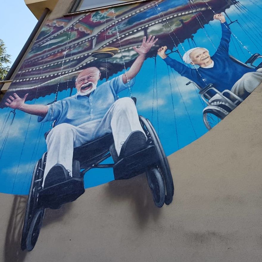 A joyful and heartwarming 3D mural by Cosimo Cheone Caiffa featuring two elderly individuals in wheelchairs enjoying a ride on a colorful swing carousel. The man in the foreground throws his arms up in delight, while the woman behind him holds onto the swing, smiling widely. The vibrant blue sky and the dynamic motion of the carousel create an uplifting scene that celebrates freedom and happiness. The mural’s incredible attention to detail and vibrant colors bring the moment to life, inspiring a sense of positivity and joy.