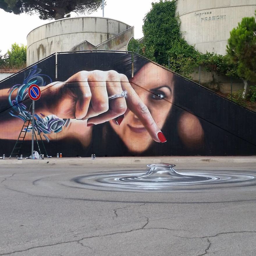 A mesmerizing 3D mural by Cosimo Cheone Caiffa, depicting a woman with a focused expression as she extends her hand to touch the pavement. Her painted finger appears to create ripples on the ground, seamlessly blending the mural with the physical street to produce a stunning illusion. The intricate details in her hand, including a shiny ring and vivid red nails, enhance the realism. The swirling water effect on the pavement adds depth and motion, making this artwork a perfect example of Cheone's skill in interactive and immersive street art.