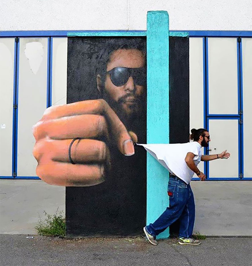 A playful 3D mural by Cosimo Cheone Caiffa depicting a man wearing sunglasses and appearing to reach out of the wall to pinch a piece of white fabric. The illusion is enhanced by an actual person walking by, with the fabric seemingly pulling their shirt, creating an interactive and humorous scene. The mural’s realistic hand and the precise integration of the artwork with the physical environment highlight Cheone’s talent for blending art and reality, making the piece both engaging and memorable.