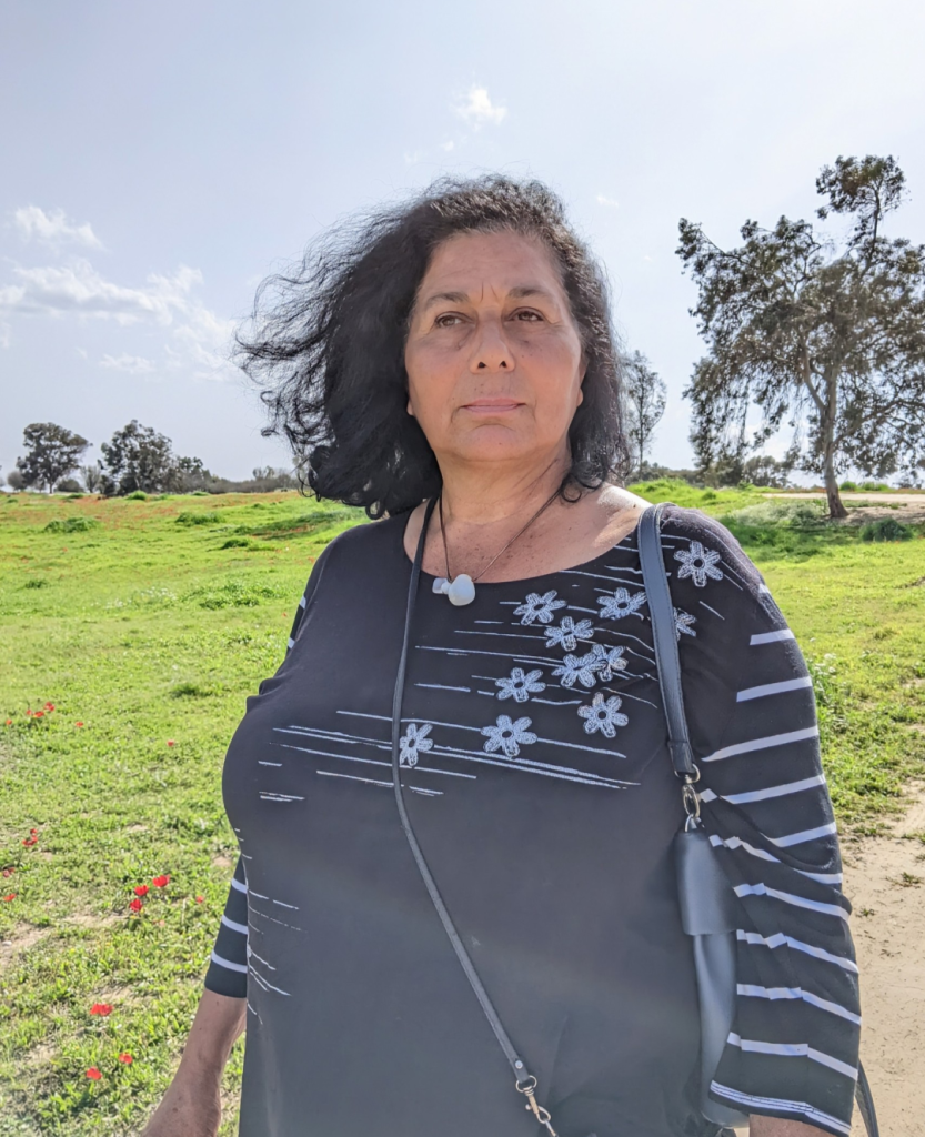 Photo of my mom at a green field with lots of red flowers and the background she is looking to the side and her dark short hair is blowing in the wind and she's wearing a black top with white flowers and a handmade necklace with white gemstone that I made for her