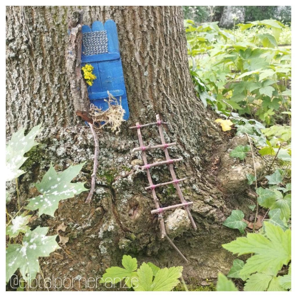 Am Fuß eines Baumstamms befindet sich eine kleine blaue Tür. Schräg darunter ist eine Leiter aus Ästen, die zusammengebunden wurden, angebracht. Ringsherum wachsen Blätter, z. B. Ahorn und Efeu.