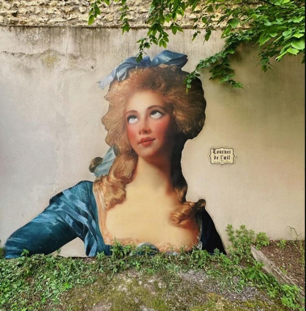 Streetart. A pretty paste-up of a lady in a rococo dress has been stuck onto a beige-colored park wall. She is wearing a large blonde wig, a blue hair bow and a blue dress with a lavish décolleté. Her cheeks are flushed and she looks up with a strangely twisted gaze. This is also the title: "Rolling eyes". Definitely a pasteup for all occasions.
Info: "L'oreille" is a French street artist who has been pasting up cities with self-designed, cut-out ears with her project "Les murs ont des oreilles" - (The walls have ears) - since around 2018. Recently, the paste-ups have been getting bigger and bigger. Whimsical works of art, full of poetry and humor.