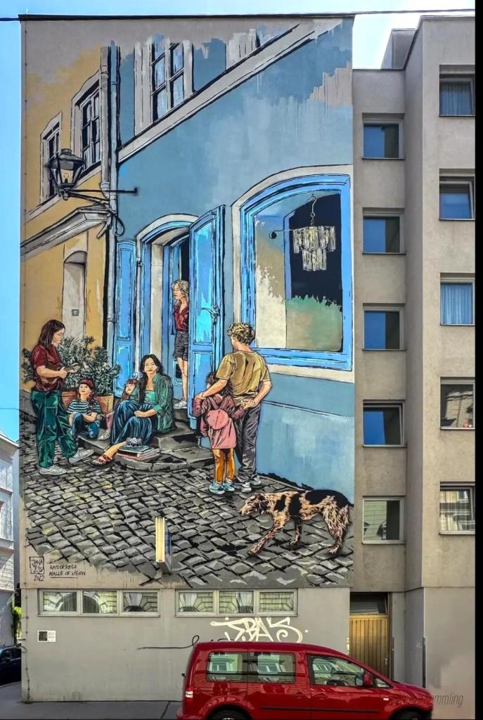 Streetartwall. A large mural (15.6 x 7.9 m) depicting a street scene was painted on the outside wall of a five-storey building. A family with neighbors in front of a blue store was painted in wonderful detail like an artistic pastel drawing. A blonde young woman in shorts stands in the doorway, a long-haired woman in a blue skirt sits on the steps in front of green plants, two little girls stand around them and a young woman stands in front of them playing the ukulele. A coat hanger with an item of clothing hangs in the shop window. A blond boy stands with his back to us, arm in arm with a smaller girl. And in the foreground of the picture, a brown dog runs across the cobblestones. What a wonderful narrative in picture form.
Info: This interpretation is based on the painting "Dorfstraße mit Leierkastenmann" by the Dutch master Adriaen van Ostade from the 17th century. The mural was financed and painted as part of the Walls of Vision project for the Kaiserschild Foundation.
