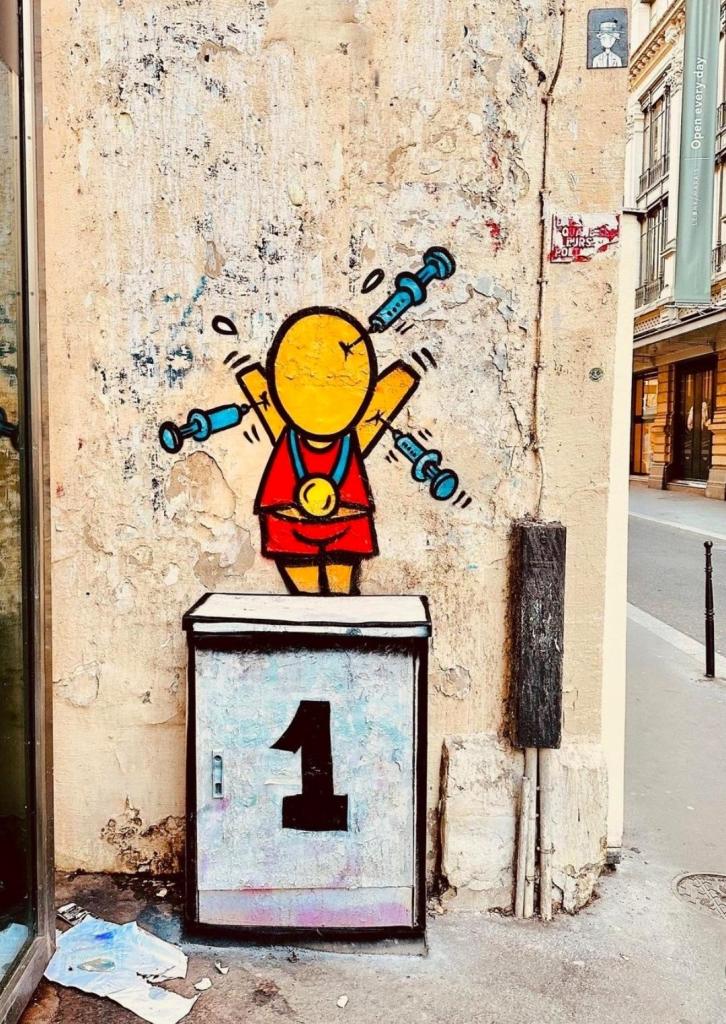 Streetartwall. On the corner of a street wall in Paris, a funny mural with a comic figure was sprayed for the Olympic Games. The figure (called Gouzou) is a small yellow man without a face, hands or feet. Wearing a red sports outfit, the Gouzou stands on a distribution box with a "1" (like a winner's podium) on it and raises his arms in the air. He is wearing a gold medal around his neck, but there are three syringes hanging from his arms and head. 