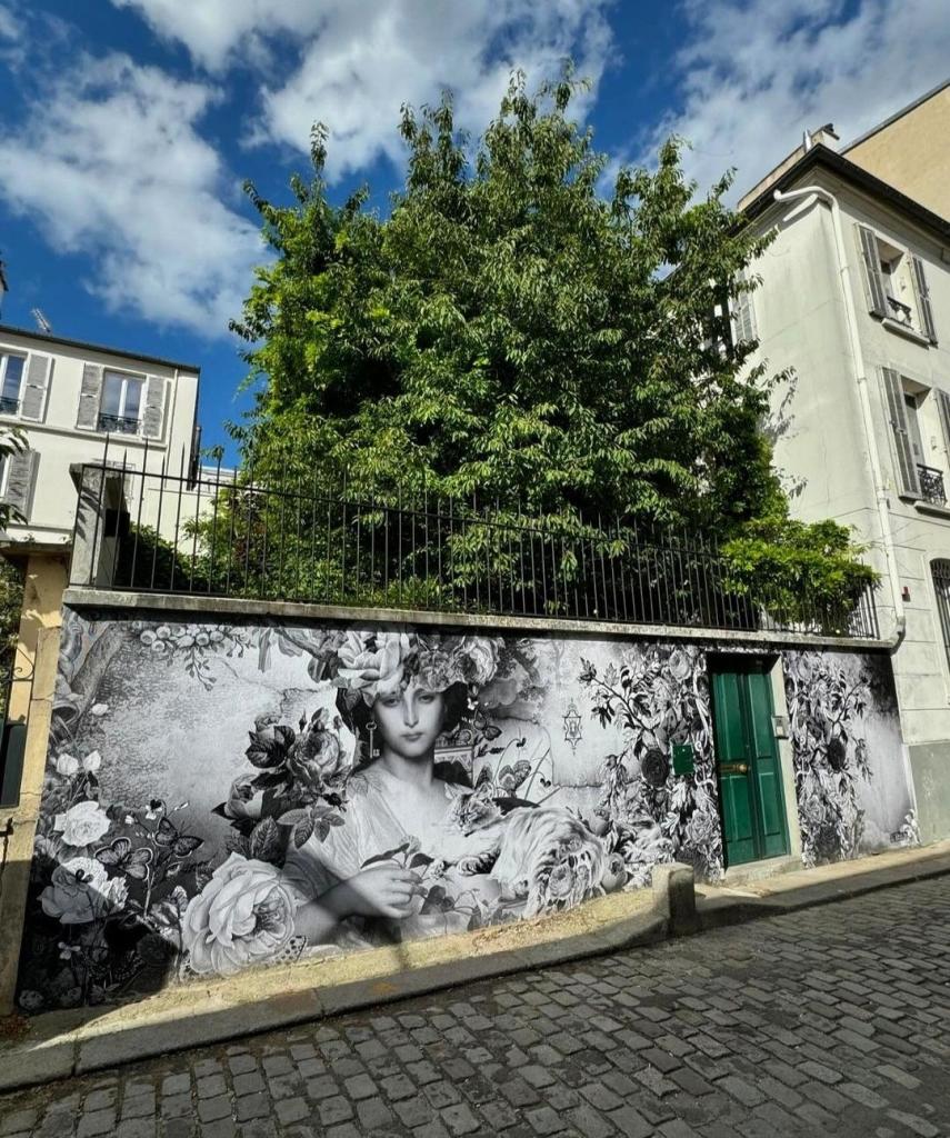 Streetartwall. A beautifully designed, black and white, poetic paste-up of a lady with a cat, surrounded by roses, was pasted on the outside wall of a long street wall. The young woman is completely surrounded by lush flora, roses and leaves fill the wall. She has a delicate face, her hair is covered by a bouquet of flowers and she is wearing a white, ornate dress. But surprisingly, the woman wears no jewelry, only an iron key as an earring. Which door does it belong to? Perhaps the sleeping Persian cat lying next to her, who seems to be the keeper of all secrets, knows. A poetic, romantic mural that inspires dreams and is absolutely Montmatre.
(The mural is interrupted by a green wooden door ( !) in the long wall. Behind the wall you can see the green foliage of a broad tree, which gives the ensemble that certain something).
Info: About the title: The painter and object artist Marcel Duchamp invented the artistic figure Rrose Sélavy. The name should be pronounced out loud: Rrose, Rose - Sélavy, C'est la vie: Pink is life.