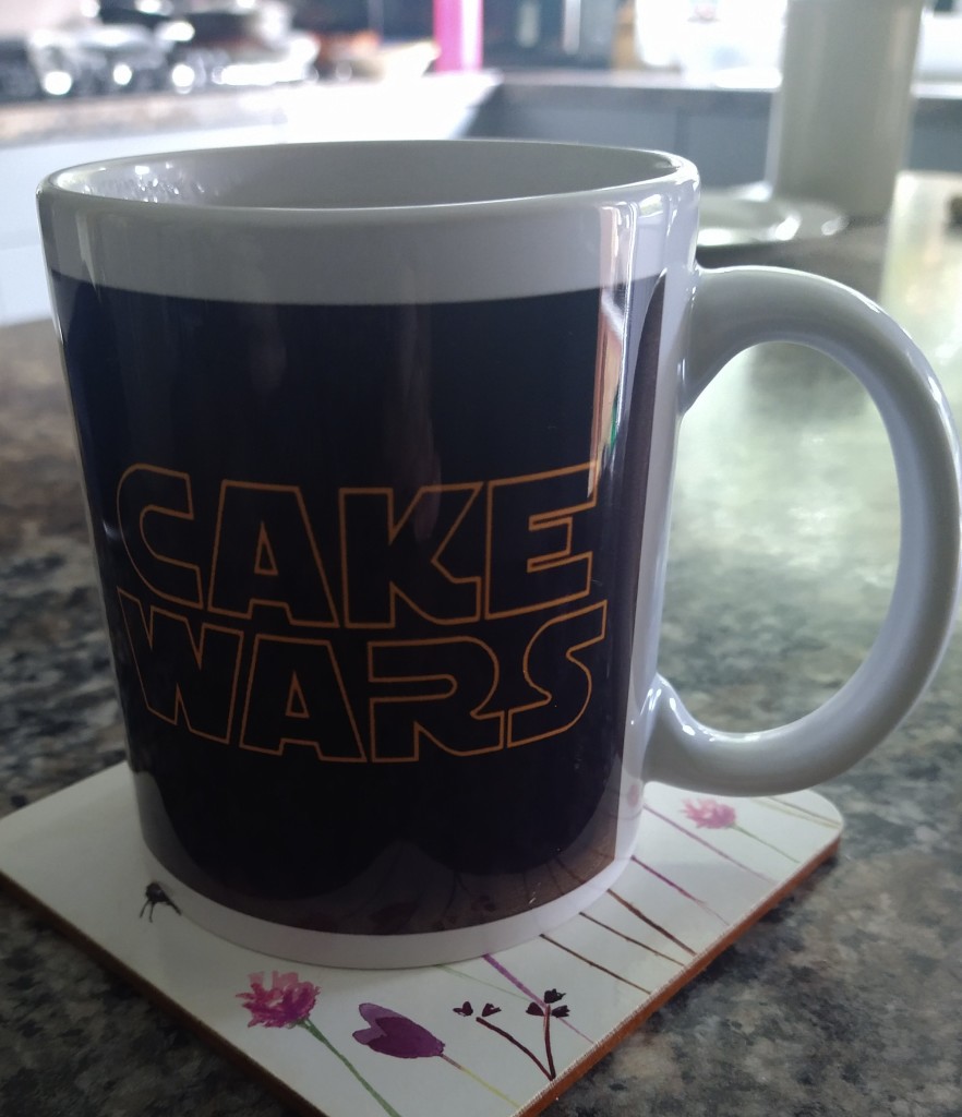 Mug of tea on a kitchen countertop. With white rim and handle, orange text saying Cake Wars on a black background 