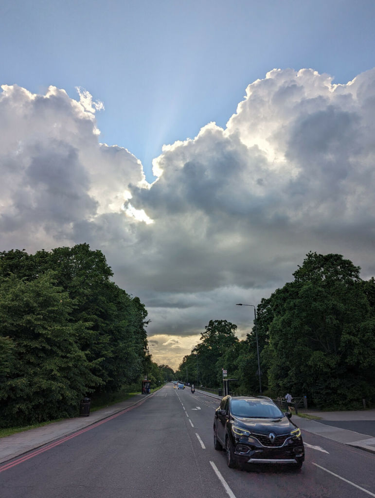 Wide-angle photo of a long straight road in the UK from just off-centre. There's very little traffic around, though there is one dark car facing the camera on the right side. On either side of the road are mounds of dark green trees. The top half of the picture is all sky and cloud. In fact the clouds continue all the way to the horizon between the banks of trees, yellowing in the distance. The clouds above give something of the sense of ragged scoops of ice-cream, somehow lit from within.