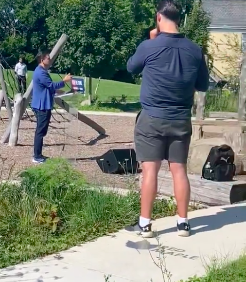 Poilievre standing in a kids playground surrounded with campaign signs. A film crew films