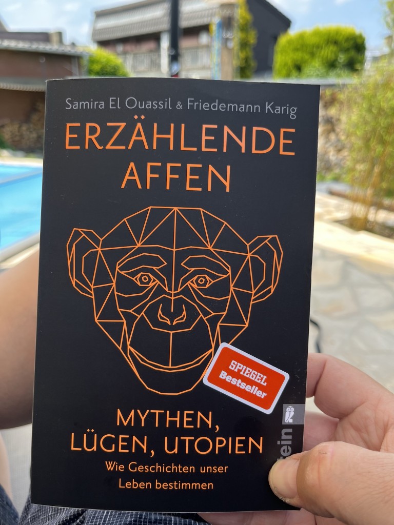 A person holding the book "Erzählende Affen" by Samira El Ouassil and Friedemann Karig. The cover features an orange geometric illustration of a monkey's face with the text "Mythen, Lügen, Utopien