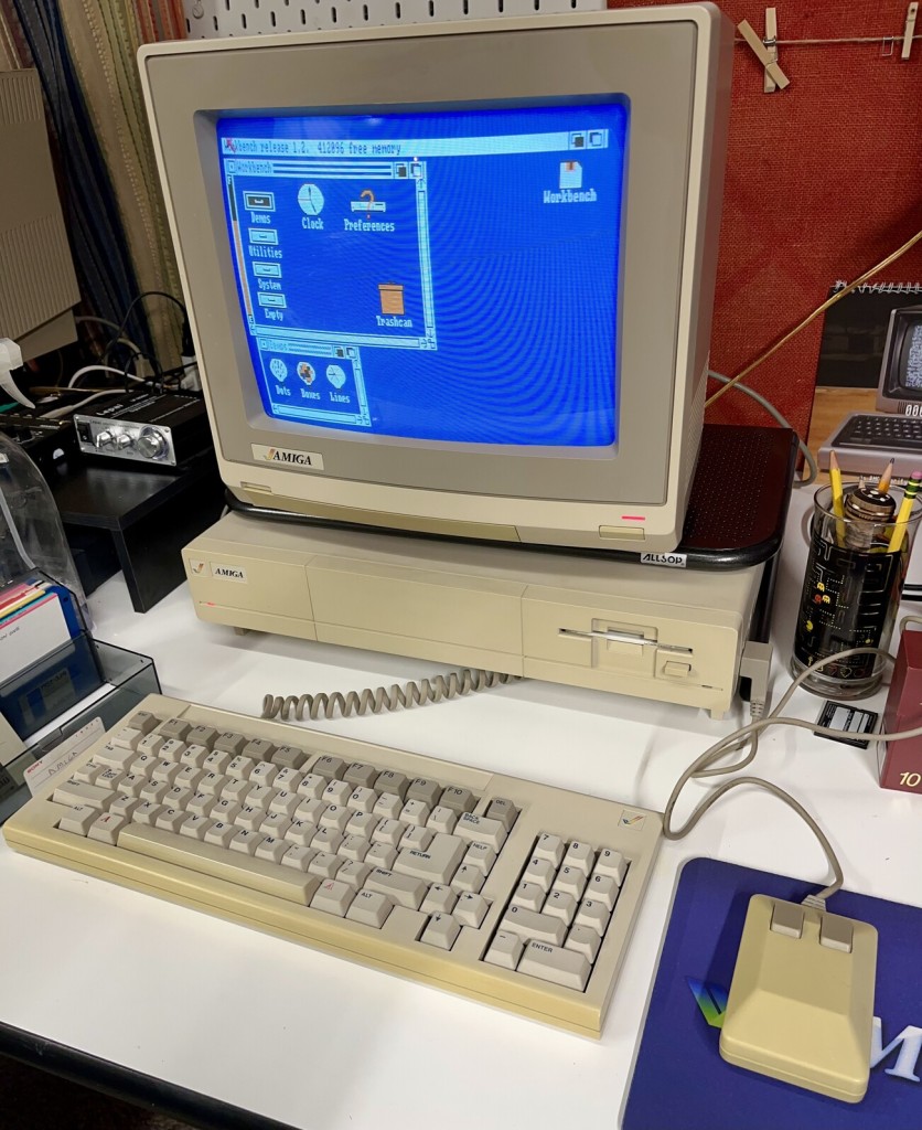 An Amiga 1000 on a desk. It’s a sorta short pizza box desktop case but with a “garage” underneath it that the keyboard fits into. The CRT monitor is showing the blue Workbench desktop with a couple windows open. 