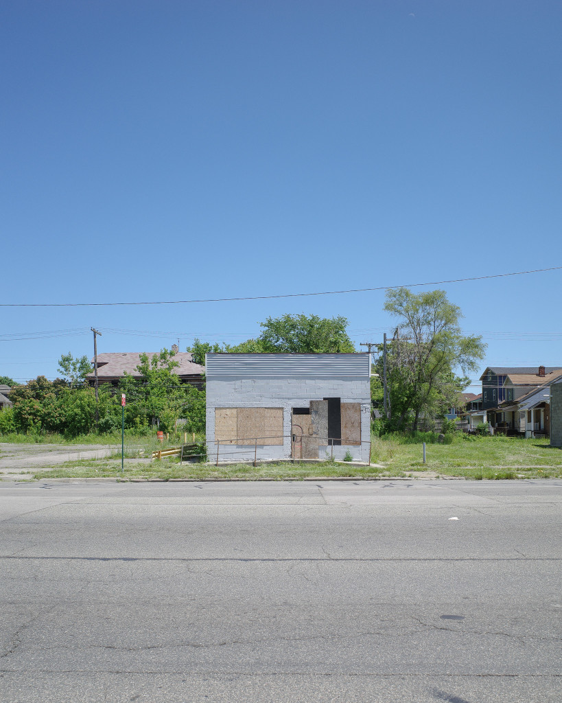 Color photograph looking across a wide gray street on a bright sunny day at a small boxy storefront building, painted gray, with boarded up windows and entrance