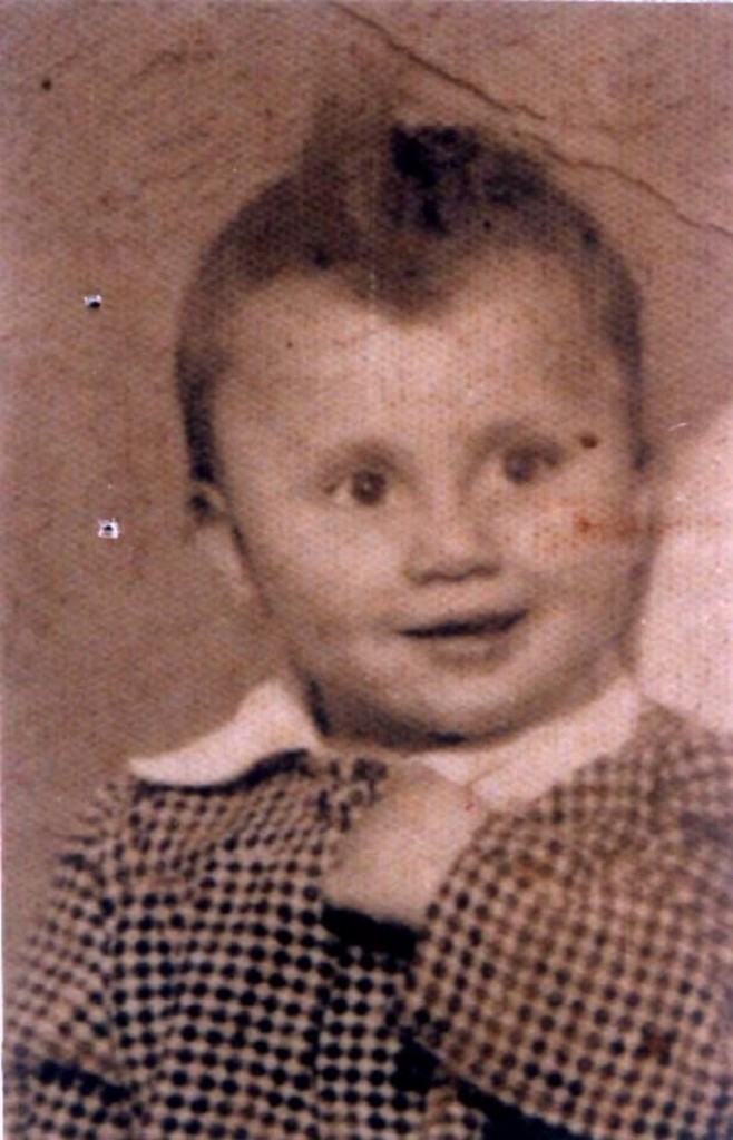 Vintage photograph of a young child with short hair, wearing a checkered outfit and a bow tie.
