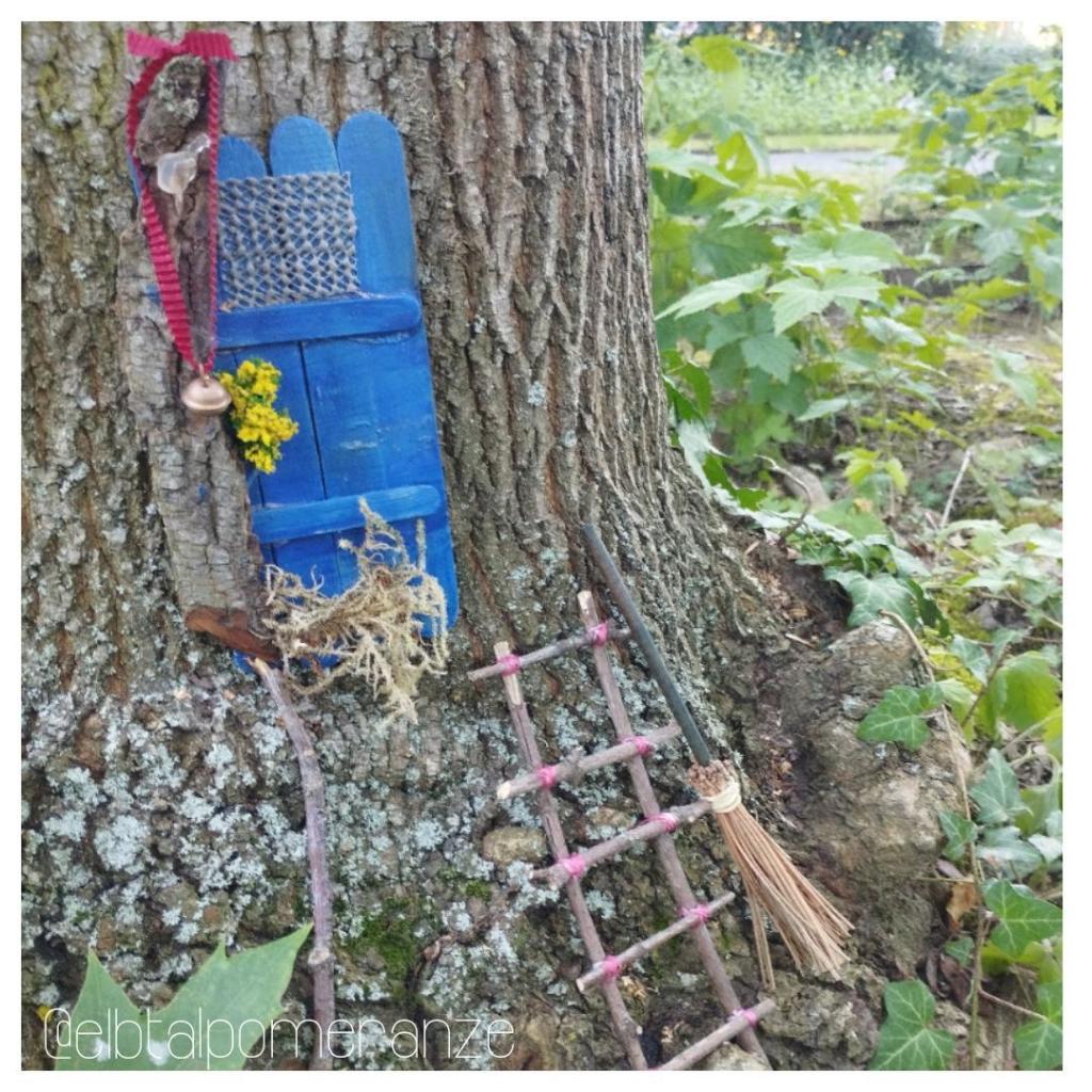 Blaue Tür am Fuß eines Baums. Daneben eine Leiter. An der Tür hängt ein Glöckchen an rotem Band. Bei der Leiter lehnt ein Reisigbesten am Baumstamm.