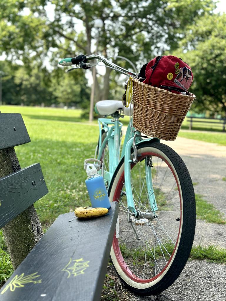 My cruiser bike, Gladys, is parked next to a low-profile park bench. There is a glass bottle full of water and a ripe banana placed at the end of the bench. Behind her, a bright sunny day at the park is visible. 