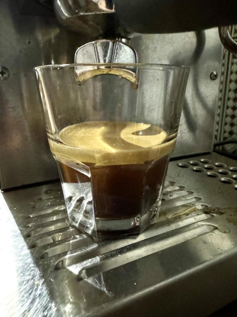 Delicious coffee, in a glass, underneath the espresso machine group head. There is a deliciously thick crema layer on top of the shot