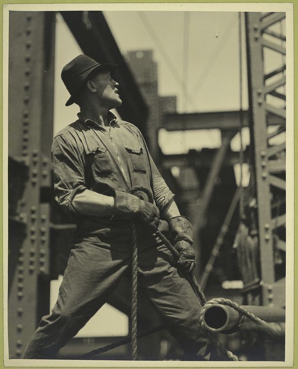  The image portrays a man in the midst of a construction scene, standing on top of what appears to be a steel structure. He is wearing a dark hat and overalls, with a belt and gloves that suggest manual labor. His gaze is directed upwards, possibly indicating that he's operating machinery or overseeing work being done above him. The background suggests an industrial setting, likely a bridge construction site. The photo has a vintage quality to it, which may be indicated by the style of clothing, the presence of a watermark or text overlay that reads "Lewis Hine," and the overall graininess of the image.