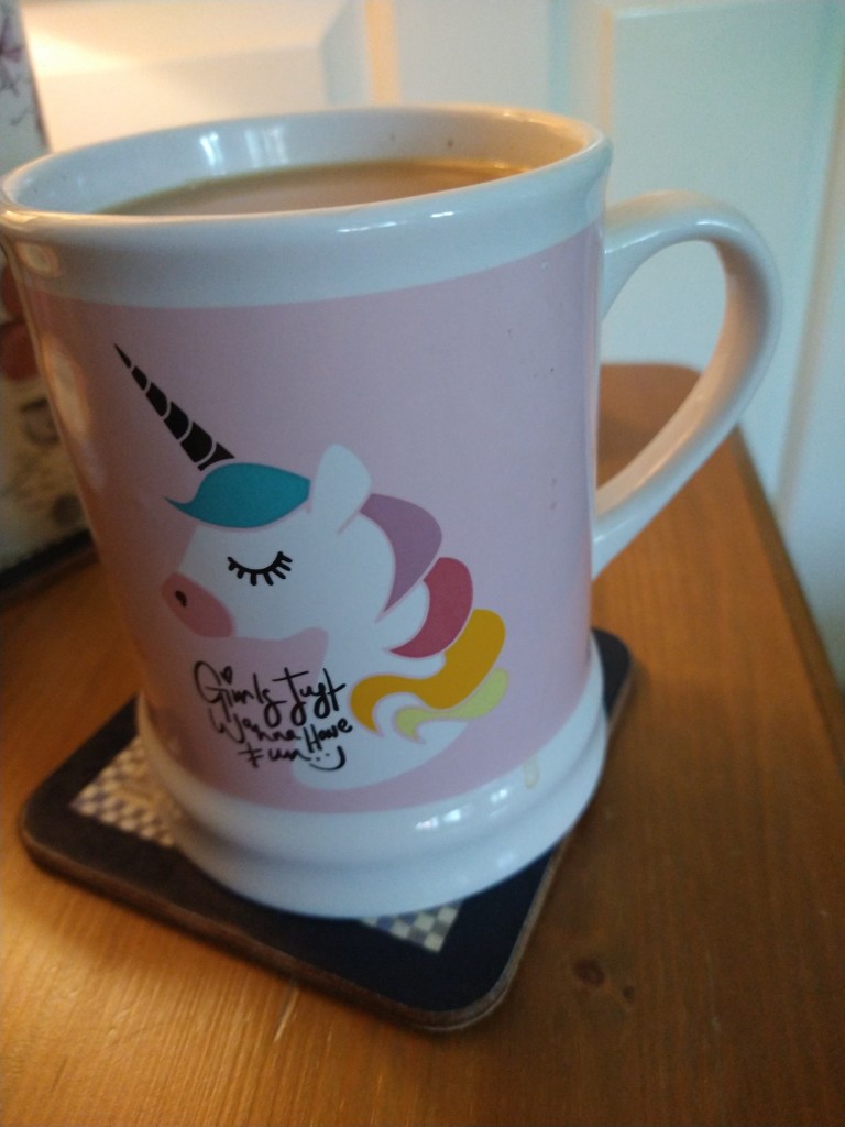 Large tankard shaped mug of tea on a bedside cabinet.  It has a pink background and a unicorn head with a black horn and multi coloured mane. 