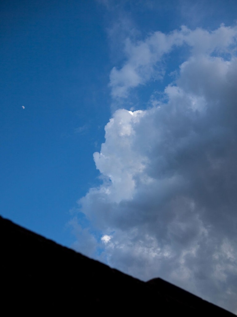 Photo with translucent-seeming clouds piling up on the right-hand side, white in places but mostly grey. Most of the rest of the picture is blue sky, shading to deep blue in the top-left corner. The moon, appearing very small, is on the left. The blurry silhouette of a building jags across the bottom-left corner.