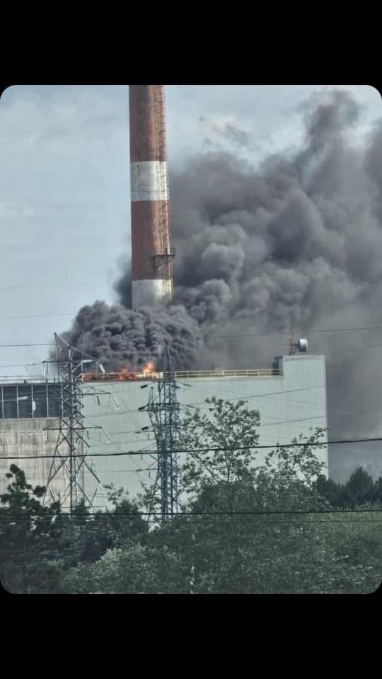 Grey black smoke billows out of a square-shaped building. Flames can be seen licking off the roof. A giant red/white striped smokestack points up like a middle finger. 