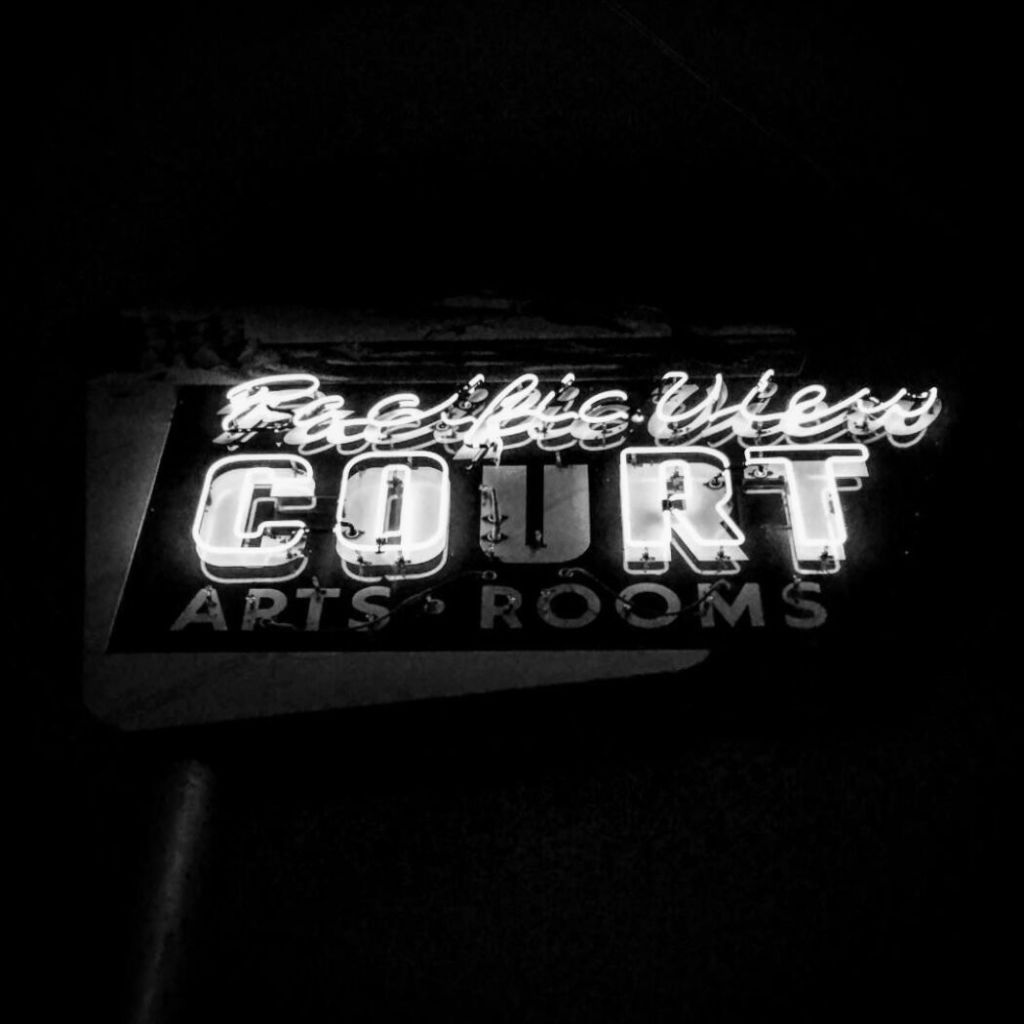 Black and white image of a neon sign that says Pacific View Court with the U burnt out or simply not illuminated. Below, in plain paint, it says apts &amp; rooms