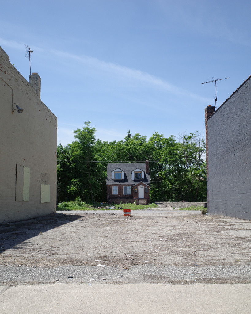 Color photograph looking across an empty concrete lot between two buildings with blank walls at a cute little brick house across the street on the other side of the bloc. The house is nestled in among some trees, looks like it has recently repaired windows and doors. In the middle of the concrete lot there's an orange barrel turned upside down and stuck in a hole