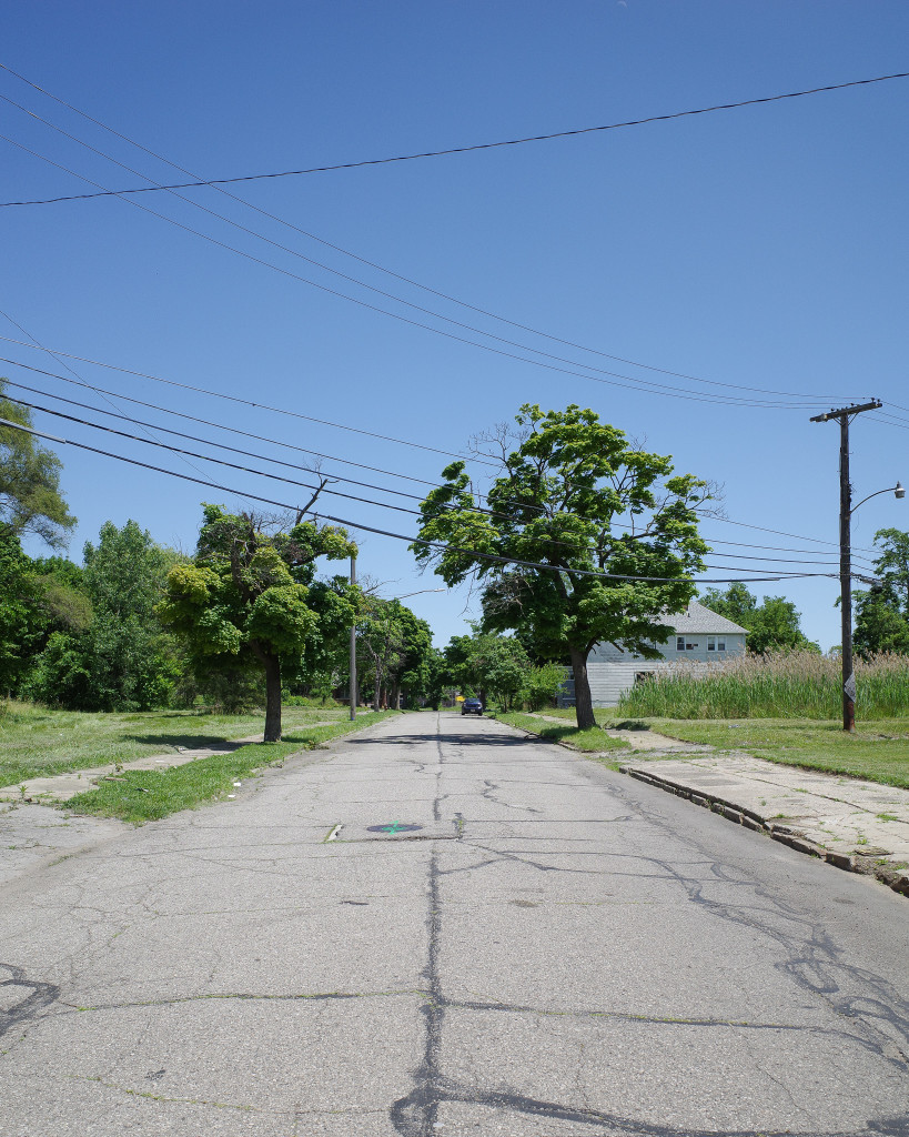 Color photograph looking down a street on a bright sunny day, mostly empty lots on either side with two trees a bit down the way casting very dark shadows, thick power lines strung across the street. There's a car parked on the street further down the block in front of a large white house