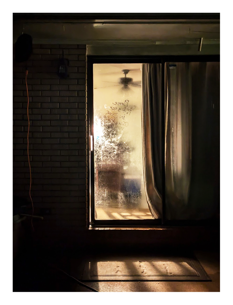 night. a sliding door has condensation, obscuring the interior of a house. a band of light illuminates a rug and the cement porch in the foreground.